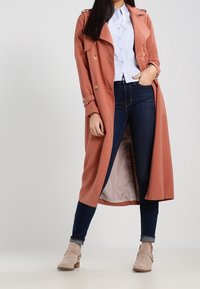 Trinchera de color óxido con botones en tono dorado, combinada con una camisa azul clara, jeans de mezclilla oscuros y botas de tobillo beige. Textura suave.