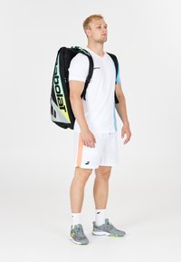 Atlet med hvid sportstøj med blå og orange detaljer står med en stor sort og neongrøn Babolat tennis taske på ryggen.