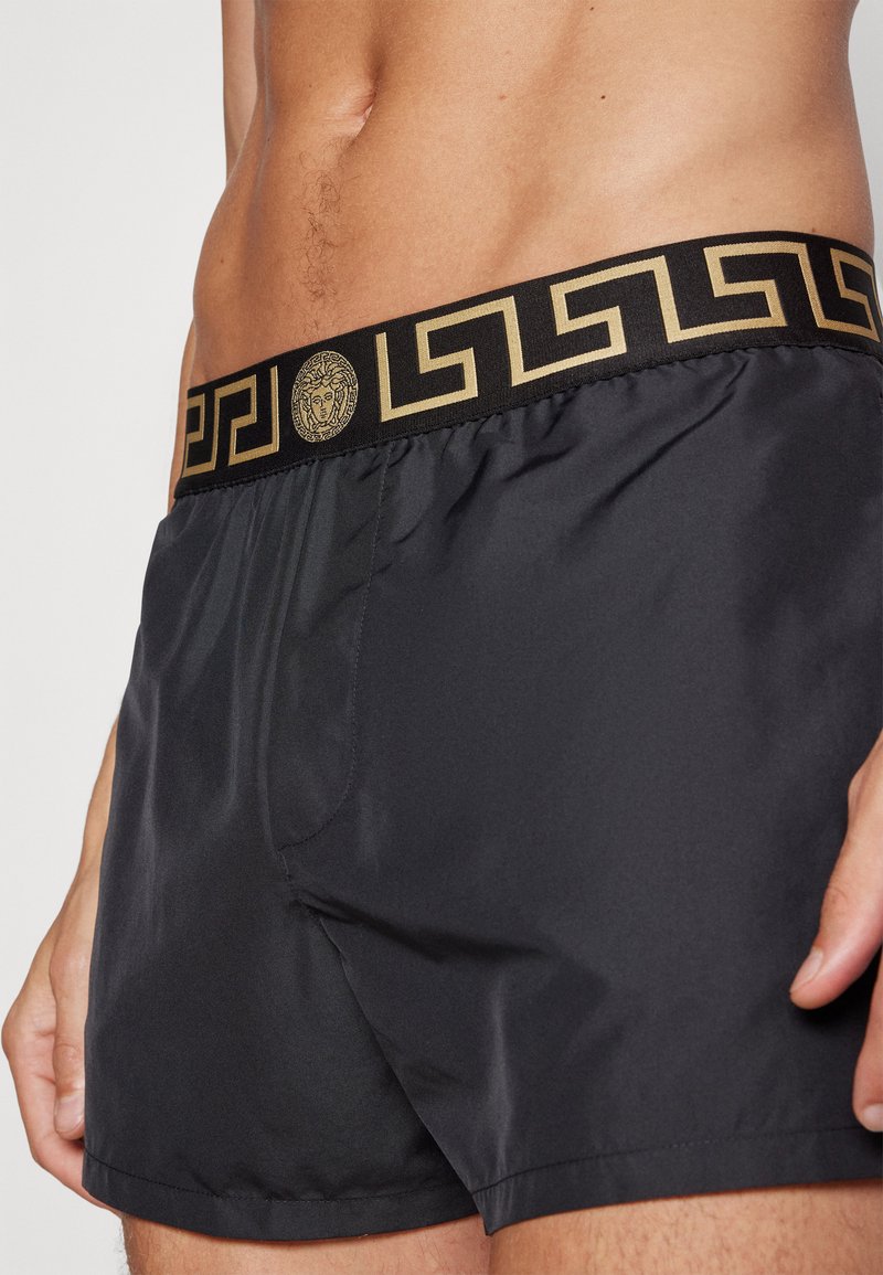 Pantaloni da nuoto neri con un elastico in vita a contrasto dorato decorato con un logo, realizzati in materiale liscio e leggero.