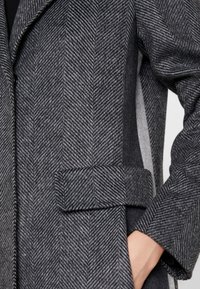 Šedý kabát v herringbone vzoru se stahovací pasem, texturovanou vlněnou látkou a kontrastním panelem v světle šedé barvě po stranách.