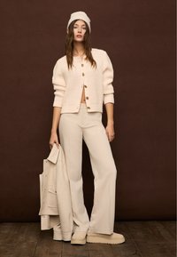 Kobieta w kremowym dzianinowym kardiganie, beżowych szerokich spodniach i czapce, trzymająca dopasowany płaszcz, stojąca na drewnianej podłodze z brązowym tłem.