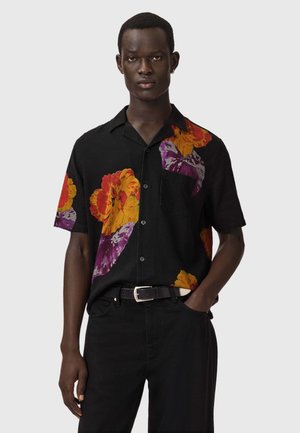 Ung mand iført en sort kortærmet skjorte med store orange og lilla blomsterprint, sorte bukser og sort bælte, stående mod en grå baggrund.