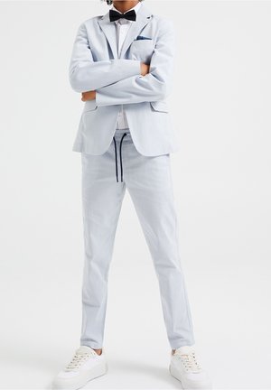 Person står med armene krydsede iført lysegrå blazer og bukser, hvid skjorte, sort butterfly og hvide sneakers på ensfarvet baggrund.