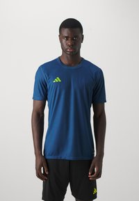 Blue athletic jersey with short sleeves, featuring a green Adidas logo on the chest. Worn with black shorts, smooth texture, and fitted design.