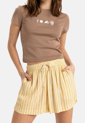 Kvinde iført brun kortærmet crop top med hvide ørken- og bølgeikoner og gule stribede shorts med snor og lommer.