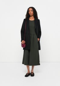 Svart ullkappa, grön midiklänning med samlad midja, maroon axelväska och svarta strukturerade skor med utskärningar. Platt bakgrund.