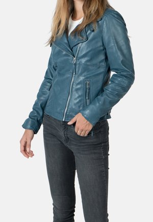 Mujer con chaqueta de cuero color verde azulado con cremallera asimétrica y vaqueros negros, de pie frente a un fondo claro y liso.
