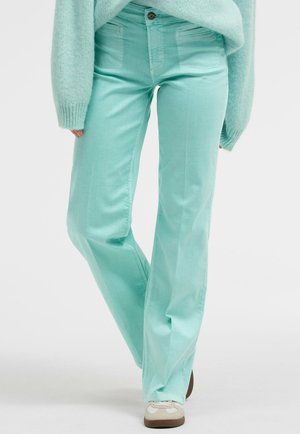 Person trägt mintgrüne gerade geschnittene Hosen, einen passenden flauschigen Pullover und weiße Turnschuhe mit braunen Sohlen, steht mit einem überkreuzten Bein.