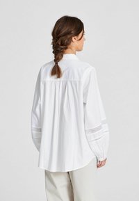 Blusa bianca a maniche lunghe con vestibilità comoda, caratterizzata da un bordo in pizzo ricamato sulle maniche e un design pieghettato sul retro. Tessuto con texture liscia.