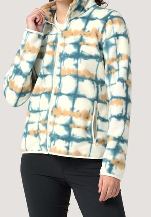 Kobieta ubrana w polarową kurtkę na zamek z niebiesko-beżowym wzorem tie-dye i zasuwanymi kieszeniami bocznymi, w parze z czarnymi spodniami.