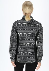 Woman with blonde hair tied in a ponytail wearing a black and white patterned long-sleeve jacket, standing facing away against a white background.