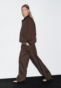 Blonde Person, die in einer gesteppten braunen Jacke, lockeren braunen Hosen und schwarzen Sandalen vor einem schlichten weißen und schwarzen Hintergrund geht.