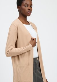 Beige cardigan met lange mouwen, open voorkant en een gestructureerde afwerking. Heeft een zak aan de voorkant en wordt gedragen over een witte top en grijze broek.