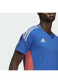 Modré sportovní tričko s V-výstřihem, s červenými akcenty po stranách a se třemi bílými pruhy na ramenou. Vyrobeno z texturované tkaniny.