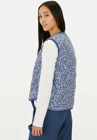 Quilted blue vest with white floral pattern, rounded neck, and armholes. Paired with a long-sleeve white shirt and denim pants.