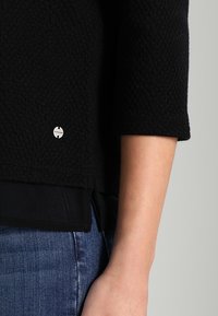 Sort struktureret sweater med trekvartærmer, der har et sølvfarvet brandlogo ved kanten, lagdelt over en sort bluse og parret med blå jeans.