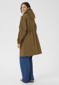 Bruine hoodie-jack met een taille die is aangesnoerd, lange mouwen en elastische manchetten. Draagt over wijde blauwe jeans. Glad, waterafstotend materiaal.
