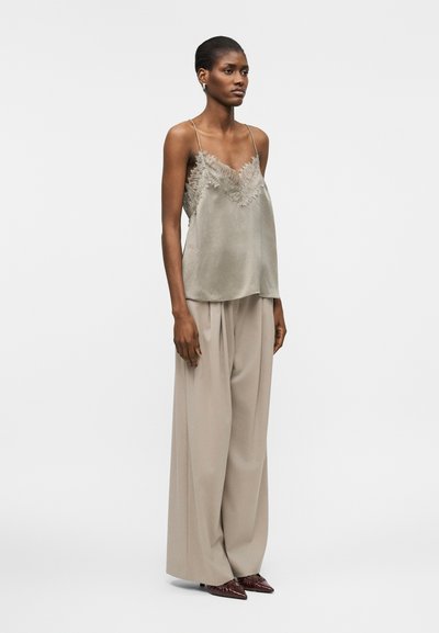 Femeie purtând o camisolă verde deschis cu dantelă, pantaloni largi bej și pantofi maro cu vârful ascuțit, stând pe un fundal alb simplu.