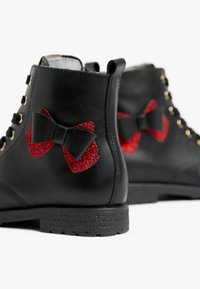 Schwarze Leder-Stiefeletten haben rote Glitzer-Schleifen an den Seiten, goldene Ösen und strukturierte schwarze Sohlen. Einfaches, robustes Design.