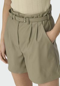 Pantalones cortos de talle alto color caqui con pliegues, un cinturón ancho con ojales de metal y hebilla, y bolsillos laterales para mayor funcionalidad.
