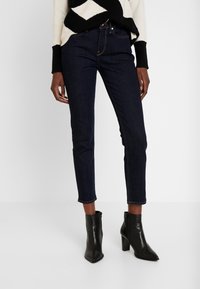 Modelka v tmavých skinny džínách a černých kotníkových botách, stylizována s vzorovaným svrškem, na jednoduchém bílém pozadí.