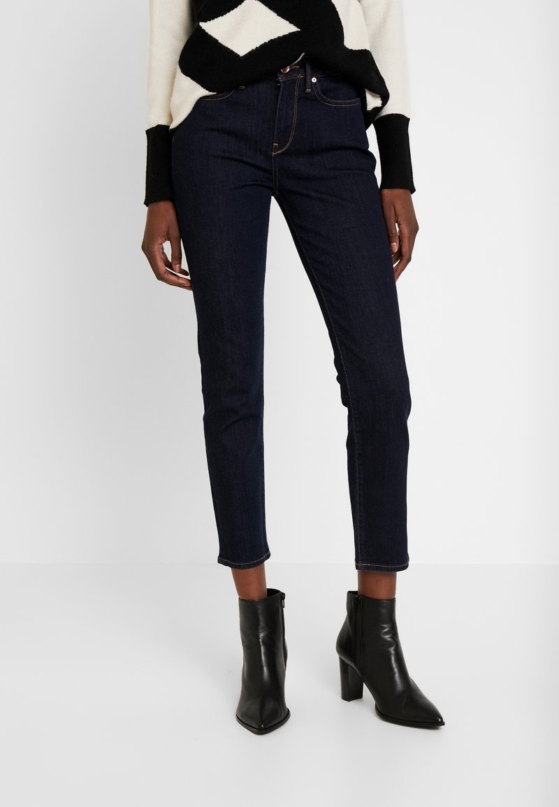 Modelka v tmavých skinny džínách a černých kotníkových botách, stylizována s vzorovaným svrškem, na jednoduchém bílém pozadí.