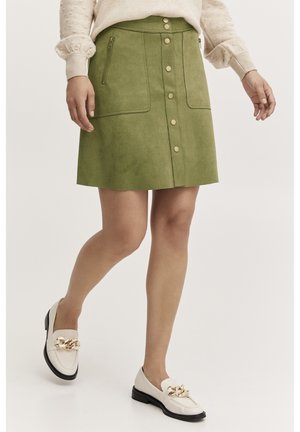 Grüner Wildleder-Minirock mit einer vorderen Knopfleiste, zwei aufgesetzten Taschen und einer A-Linie Silhouette. Kombiniert mit weißen Slippern mit goldenen Akzenten.
