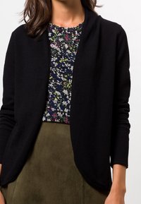 Cardigan texturé noir avec un motif en maille gaufrée, ouverture sur le devant et manches longues, porté sur un haut à imprimé floral et une jupe couleur neutre.