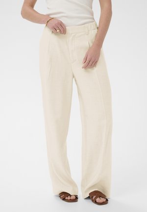 Personne portant un pantalon crème taille haute à jambes larges avec un haut blanc sans manches et des sandales brunes ouvertes aux orteils, les mains reposant près de la taille.