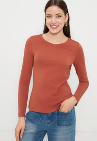 Γυναίκα που χαμογελά, φοράει μακρυμάνικο πουκάμισο σε σκουρόχρωμο πορτοκαλί και μπλε τζιν, με το ένα χέρι στην τσέπη, όρθια μπροστά σε λευκό φόντο.