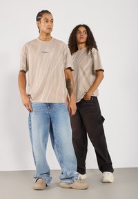 Dva modely stojí vedle sebe. Mají na sobě oversized béžové trička se šikmými pruhy, doplněná modrými a černými volnými kalhotami.