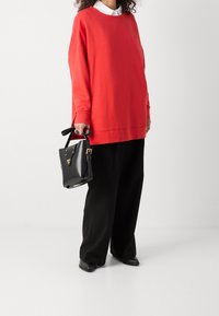 Rode oversized trui met verlaagde schouders, gecombineerd met zwarte broek met wijde pijpen. Houdt een kleine zwarte handtas met gouden accenten vast.