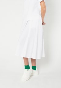 Gonna bianca plissettata in tessuto liscio, abbinata a sneakers bianche e calzini verdi, che mostrano un design pulito e minimalista.