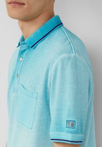 Turkusowa koszulka polo o teksturowanym splocie, z trzema guzikami na plastrze, kieszenią na lewej piersi oraz kołnierzykiem w purpurowe i niebieskie paski.