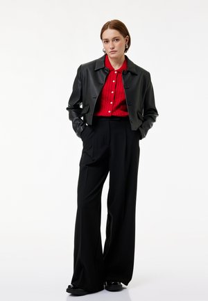 Kobieta stojąca z rękami w kieszeniach, ubrana w czarną skórzaną krótka kurtkę, czerwoną koszulę zapinaną na guziki, szerokie czarne spodnie i czarne buty.