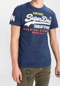 Marinblå bomulls T-shirt med rundad halsringning, med vit och röd text på framsidan och en kontrasterande vit bokstav på ärmen.