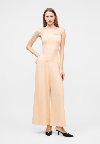 Γυναίκα με μακριά κυματιστά μαλλιά φορούσε μία μπεζ ολόσωμη φόρμα χωρίς τιράντες με φαρδιά πόδια και μαύρες μυτερές γόβες με χαμηλό τακούνι, στέκεται μπροστά σε απλό φόντο.