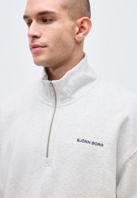 Νεαρός άνδρας με ανοιχτόχρωμο δέρμα και κοντά μαλλιά φοράει ανοιχτό γκρι πουλόβερ με φερμουάρ μέχρι το λαιμό και λογότυπο "BJÖRN BORG" στο στήθος, στραμμένος προς τα αριστερά.