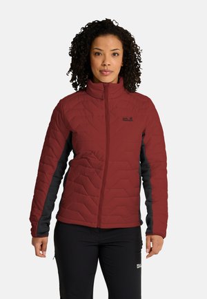 Donna con capelli ricci che indossa una giacca outdoor rossa e nera con zip e pantaloni neri, in piedi su uno sfondo semplice.