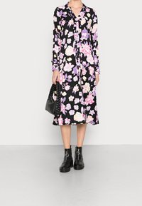 Blumiges Midi-Kleid mit schwarzem Hintergrund, verziert mit rosa und lila Blumen, langen Ärmeln, Kragen und Knopfleiste. Kombiniert mit schwarzen Stiefeln und einer Handtasche.