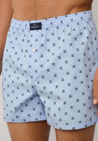 Plave kratke gaće s uzorkom koje sadrže dizajn škara u mornarsko plavoj boji. Izrađene su od lagane tkanine s elastičnom trakom u struku i prednjim zatvaračem na gumb.