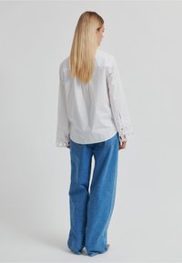 Chemise blanche à manches longues avec détail d'encoche circulaire aux poignets, associée à un jean large en denim bleu, tous deux décontractés.