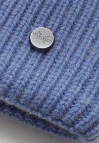 Světle modrý pletený kulich s žebrovanou strukturou, se stříbrným knoflíkem označeným "PEARL.WOOD." Materiál vypadá měkký a teplý.