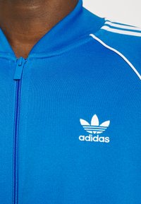 Modrá zipová bunda vyrobená z texturované látky, se bílým logem Adidas a třemi bílými pruhy na ramenou. Hladký zip.