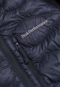 Schwarze isolierte Jacke mit gestepptem Design, Vorderreißverschluss und gesticktem "Peak Performance"-Logo auf der Brusttasche.