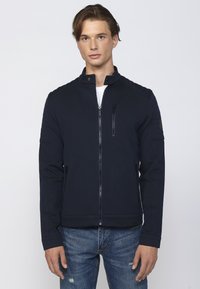 Jachetă bleumarin cu fermoar, cu guler înalt, material texturat, accente pe umeri și buzunare laterale; combinată cu blugi din denim distruși.
