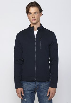 Chaqueta de punto azul marino con cremallera, cuello alto, tela texturizada, acentos en los hombros y bolsillos laterales; combinada con jeans de mezclilla desgastados.