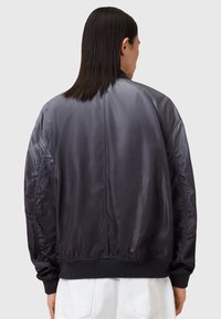 Musta satiininen bomber-takki, jossa on ribbed hihansuut ja vyötärö; erottuu sileällä pinnalla ja minimalistisella muotoilulla, ilman näkyviä logoja tai koristeita.