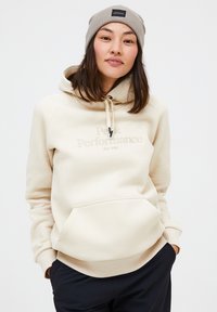 Beige fleece hættebluse med en kengurulomme, snoretræk i hætten og "Peak Performance" logo. Kombineret med mørke bukser med ribbede mansjetter.