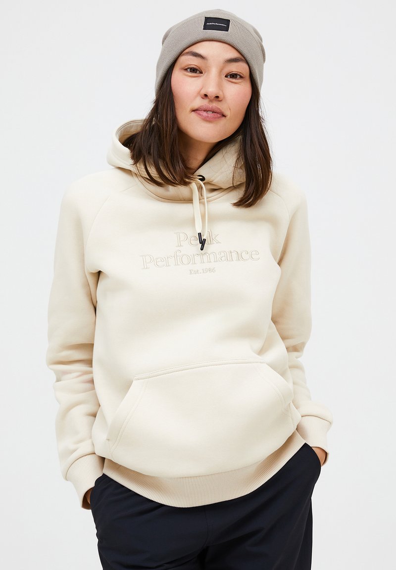 Beige fleece hættebluse med en kengurulomme, snoretræk i hætten og "Peak Performance" logo. Kombineret med mørke bukser med ribbede mansjetter.